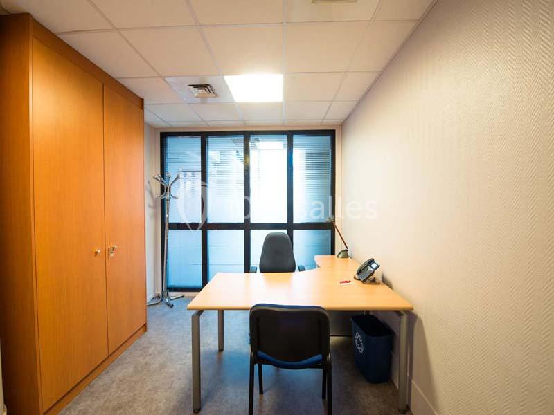 Bureau simple avec une table, deux chaises, une armoire en bois, un téléphone et une fenêtre avec stores.