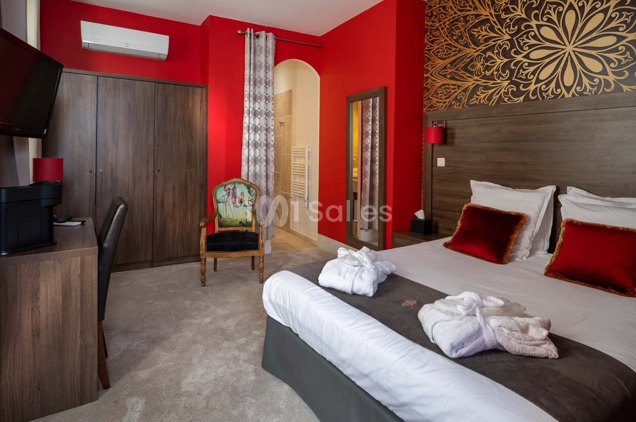 Chambre d'hôtel avec lit double, fauteuil, armoire, bureau, et salle de bain attenante, décorée en rouge et bois.