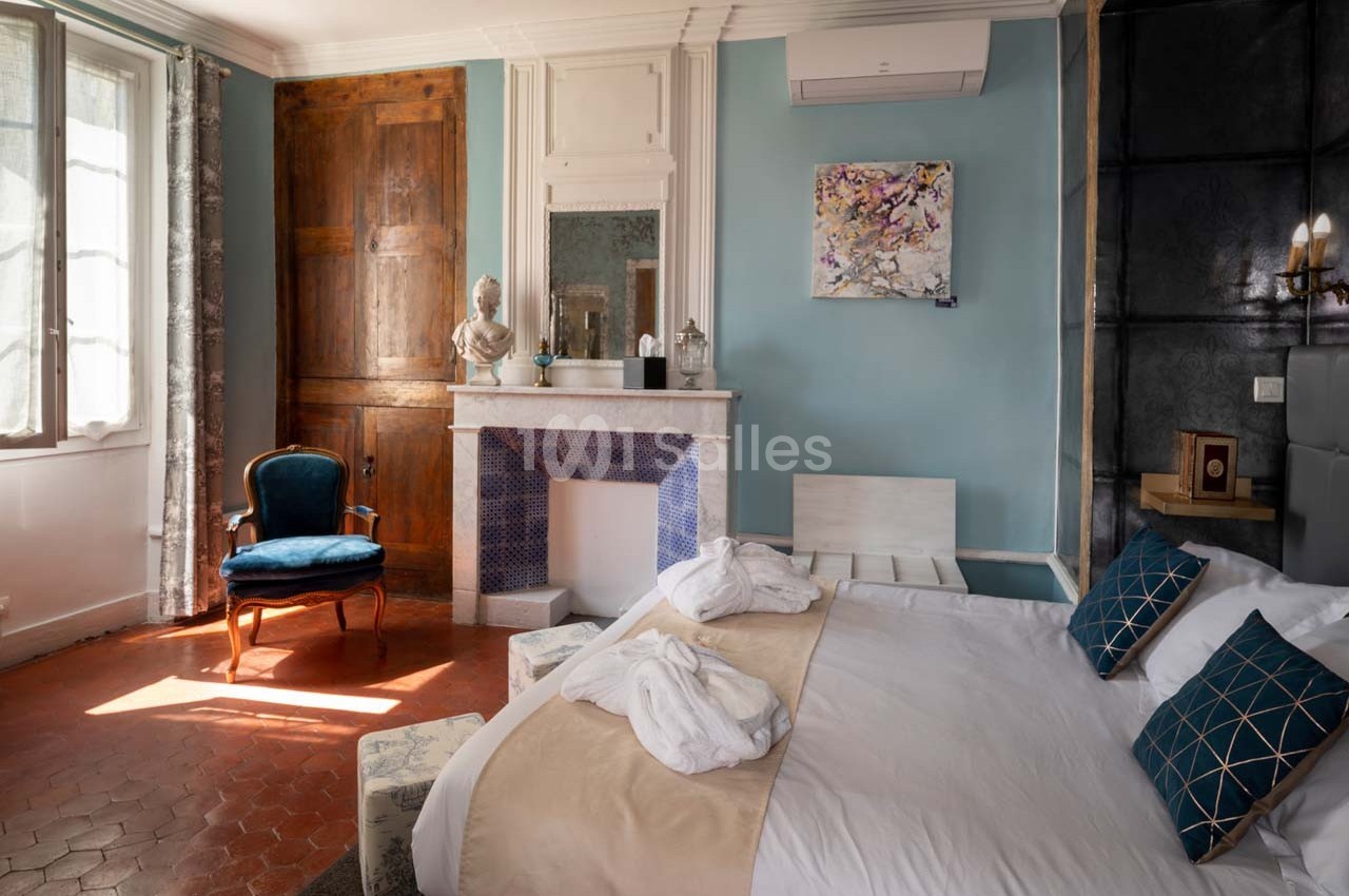 Chambre lumineuse avec lit double, fauteuil bleu, cheminée décorative et murs bleu clair.