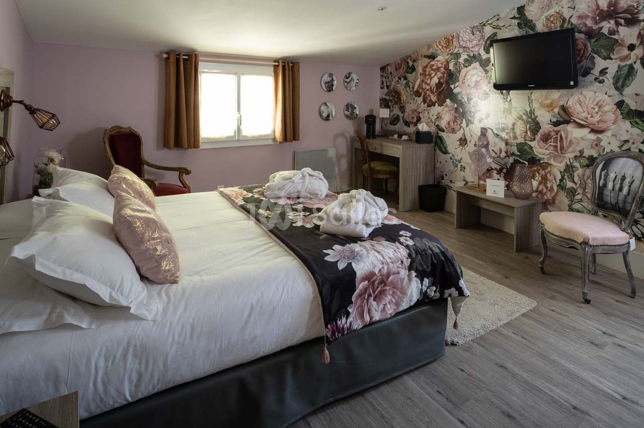 Chambre décorée avec un lit double, fauteuil, bureau, télévision murale et mur fleuri, éclairée par une fenêtre.