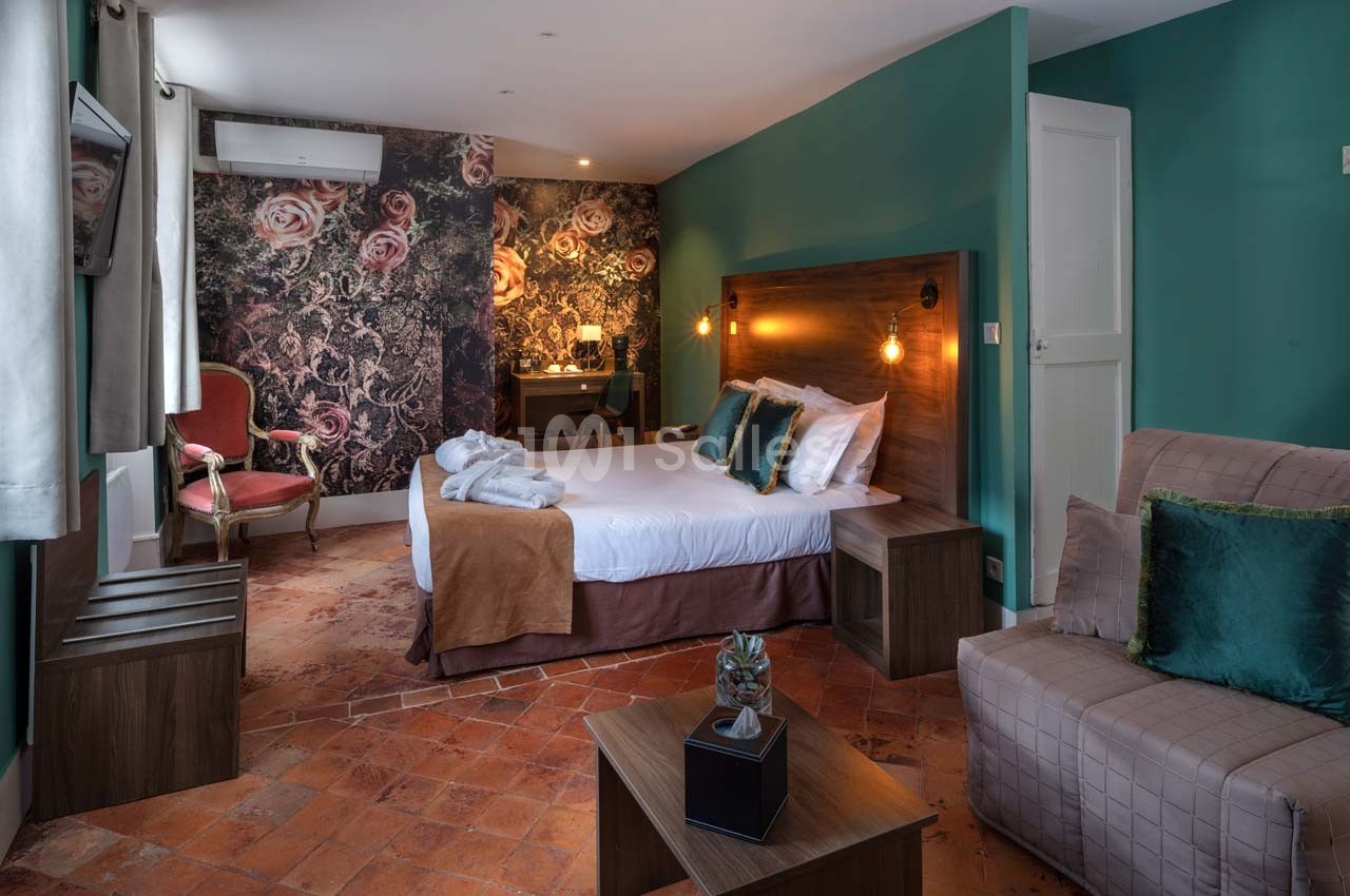 Chambre lumineuse avec lit double, fauteuils, bureau, murs décorés de motifs floraux et sol en carreaux de terre cuite.