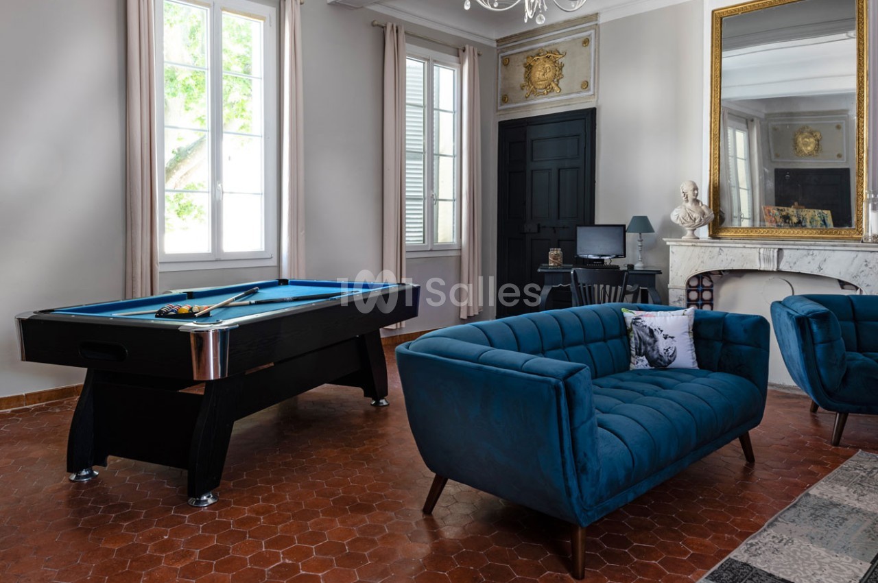 Salle de séjour lumineuse avec un billard, des fauteuils bleus, un bureau, une cheminée et un grand miroir doré.