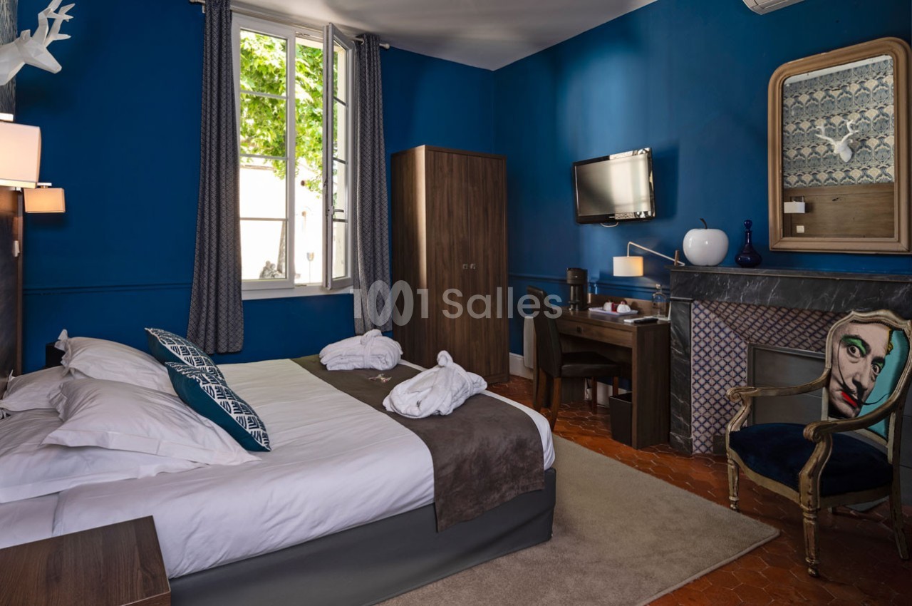 Chambre d'hôtel avec murs bleus, lit double, bureau, fauteuil décoratif et fenêtre donnant sur un arbre.