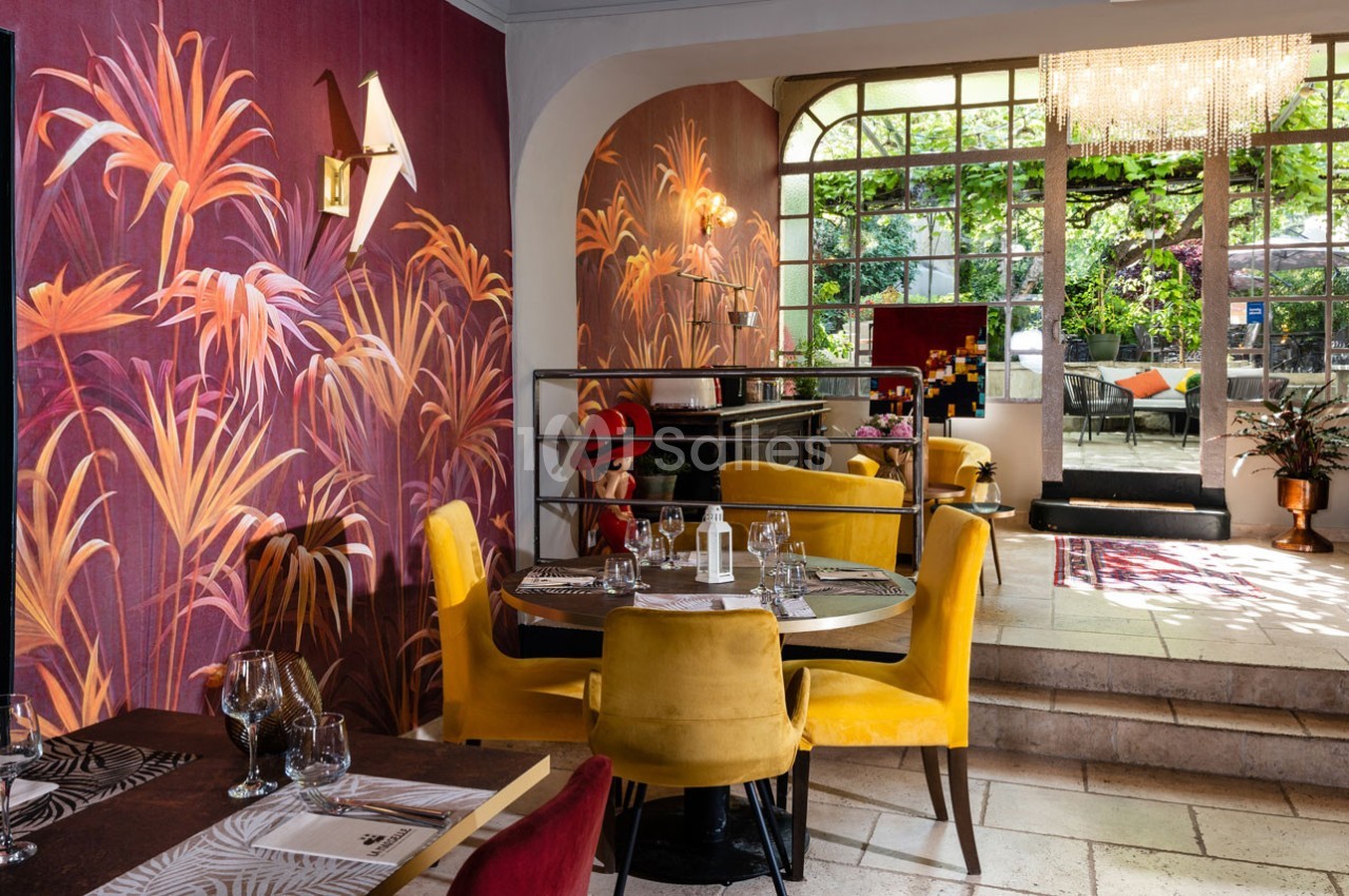 Salle de restaurant élégante avec murs ornés de motifs végétaux colorés, mobilier jaune et vue sur une terrasse verdoyante.