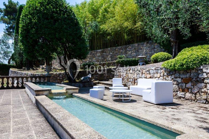 Mobilier de jardin blanc disposé près d'un bassin rectangulaire entouré de végétation et d'un mur en pierre.