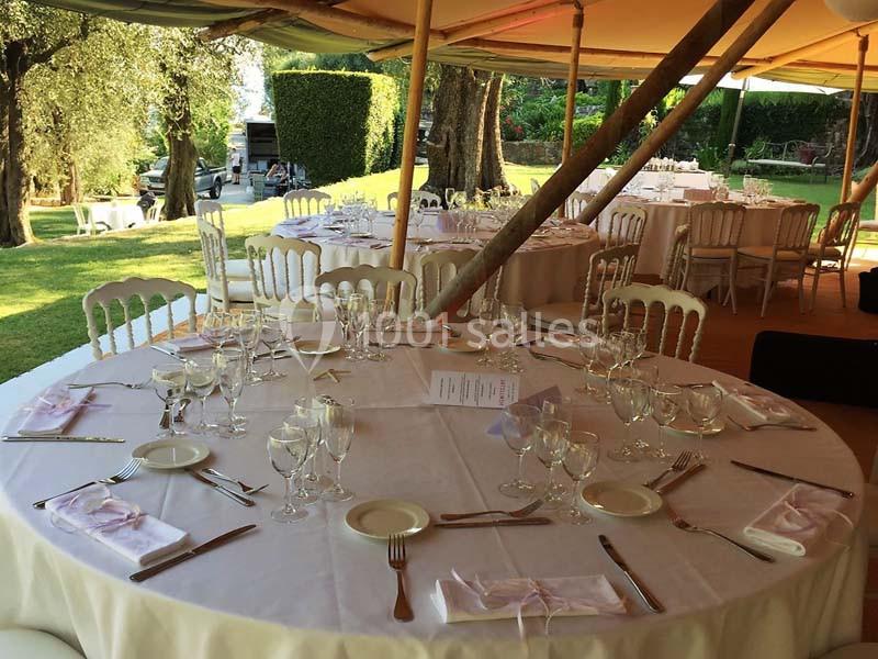 Tables rondes dressées avec nappes blanches, couverts et verres, sous une tente dans un jardin verdoyant.