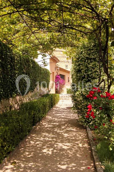 Allée ombragée bordée de haies et de fleurs colorées menant à une maison aux murs clairs sous un ciel dégagé.