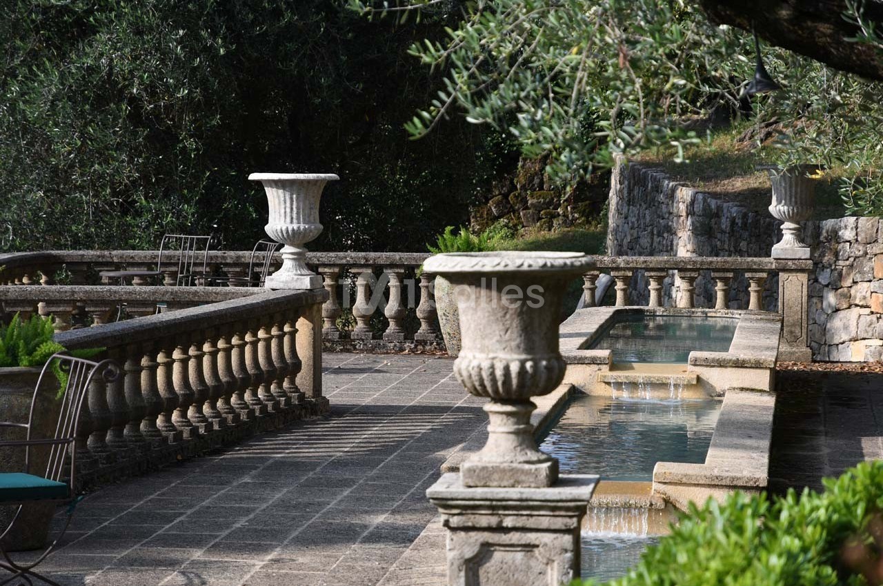Terrasse en pierre avec balustrades, fontaines en cascade et urnes décoratives entourées de végétation.