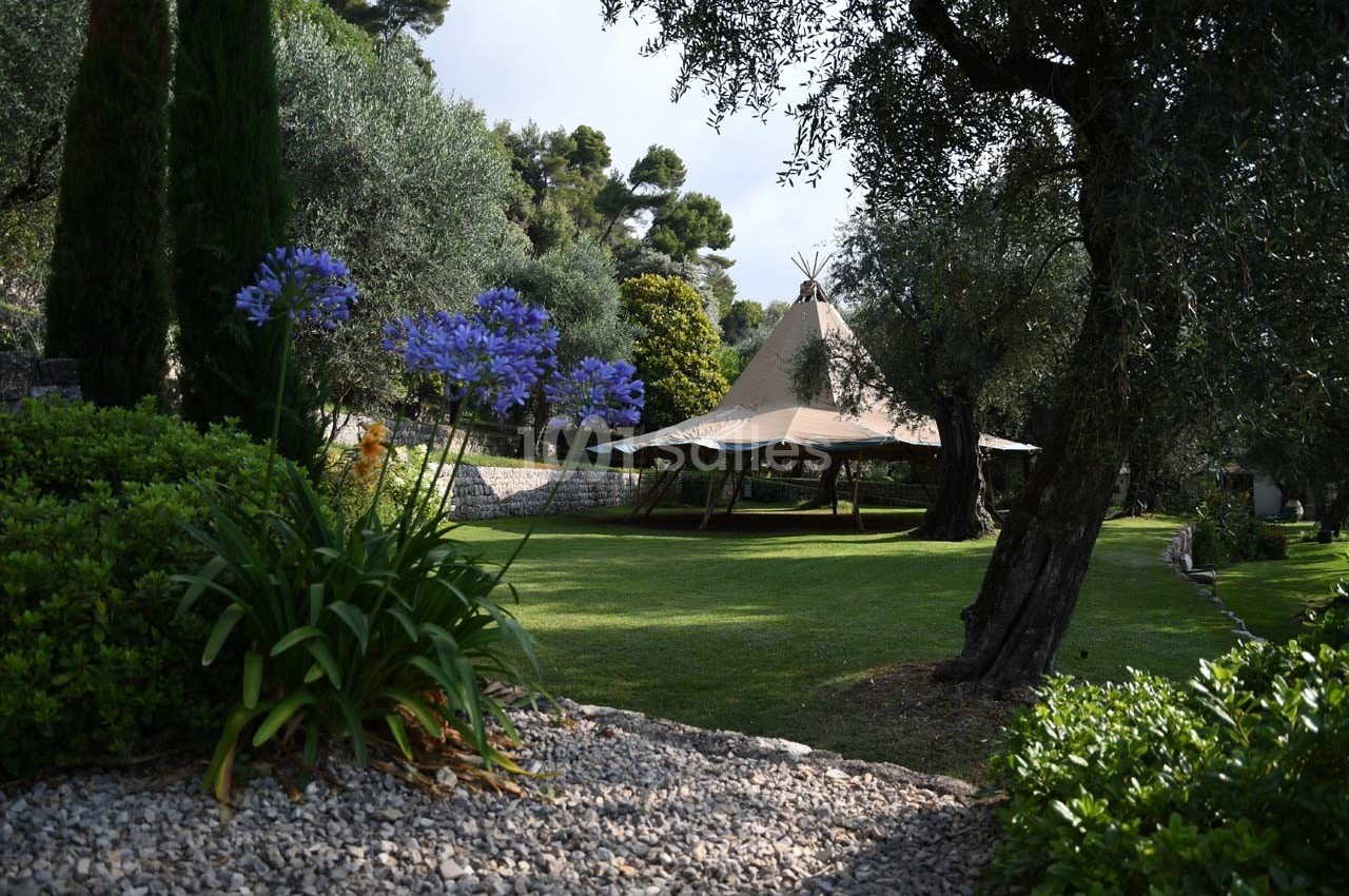 Tente beige installée dans un jardin verdoyant avec des oliviers et des fleurs violettes au premier plan.