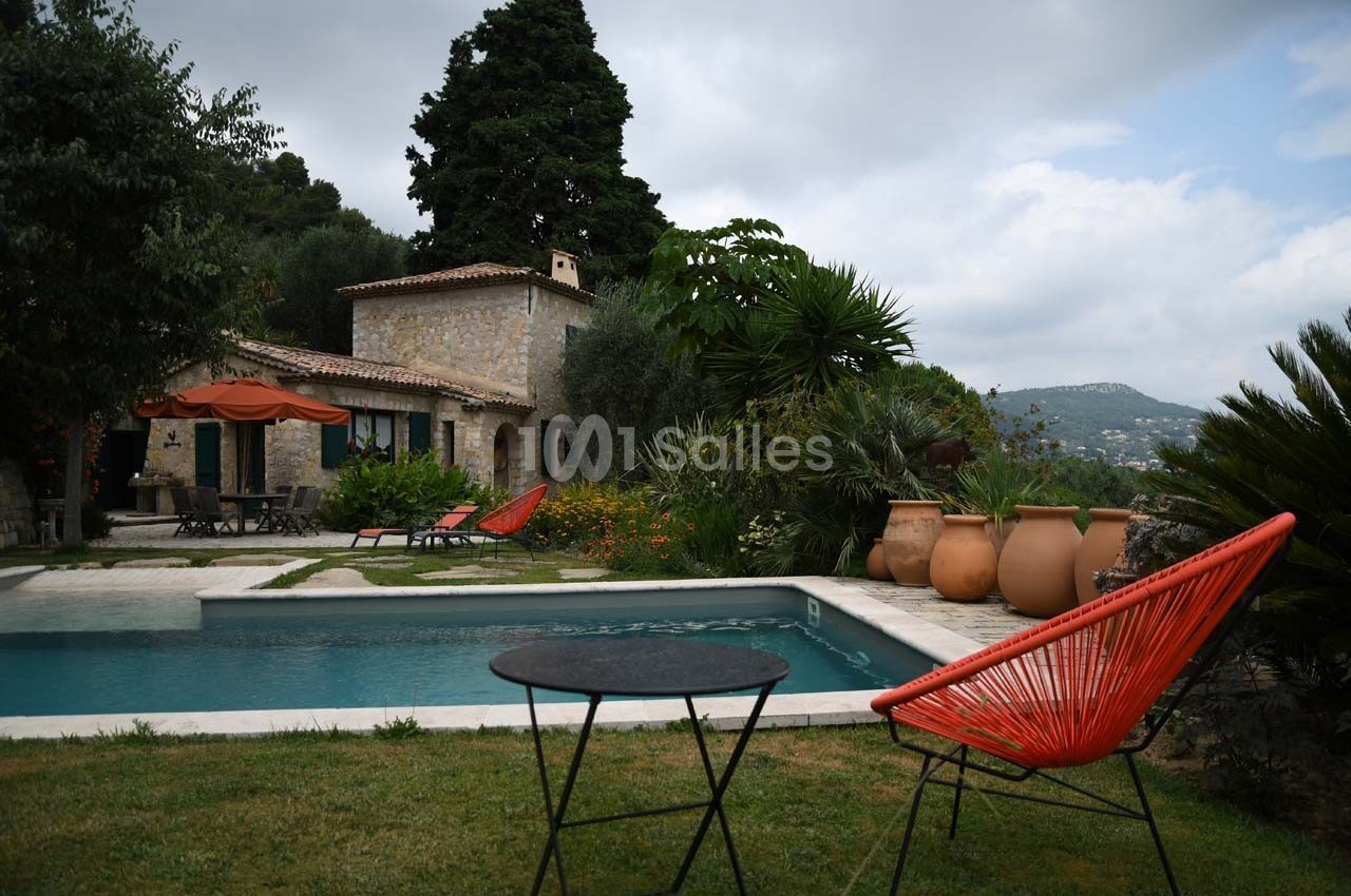 Jardin avec piscine, maison en pierre, chaises colorées et végétation méditerranéenne sous un ciel nuageux.