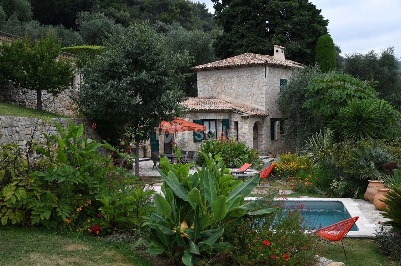 Maison en pierre entourée de végétation luxuriante avec une piscine et des chaises colorées dans un jardin.