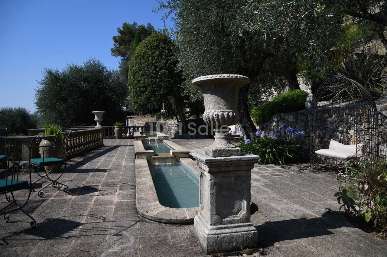 Terrasse en pierre avec bassin central, urne décorative, chaises en fer forgé et végétation méditerranéenne.