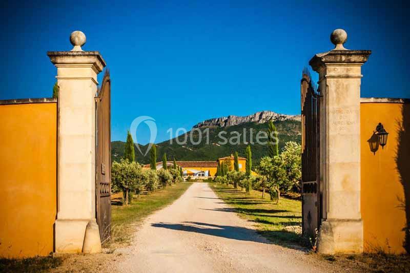 Entrée d'un domaine avec portail ouvert, chemin bordé d'oliviers menant à une maison sous un ciel bleu clair.