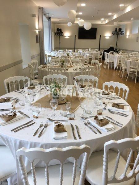 Salle de réception décorée pour un événement, tables rondes dressées avec nappes blanches, fleurs et couverts.