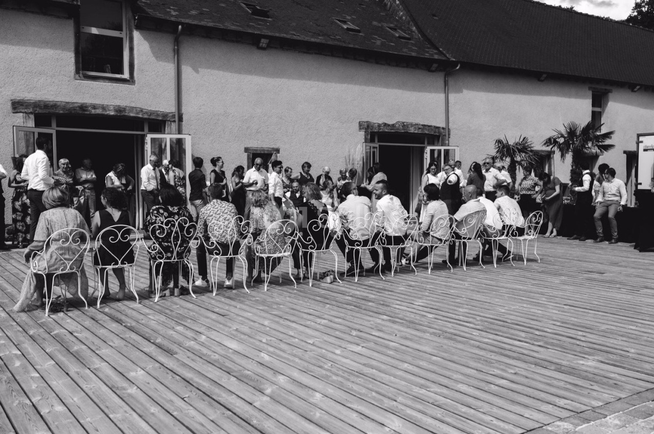 Groupe de personnes rassemblées sur une terrasse en bois devant un bâtiment, certaines assises sur des chaises en fer forgé.