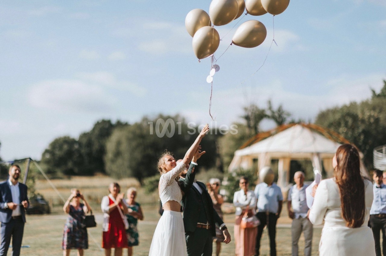 Une femme en robe blanche lâche des ballons dorés dans un jardin, entourée de personnes en tenue de cérémonie.