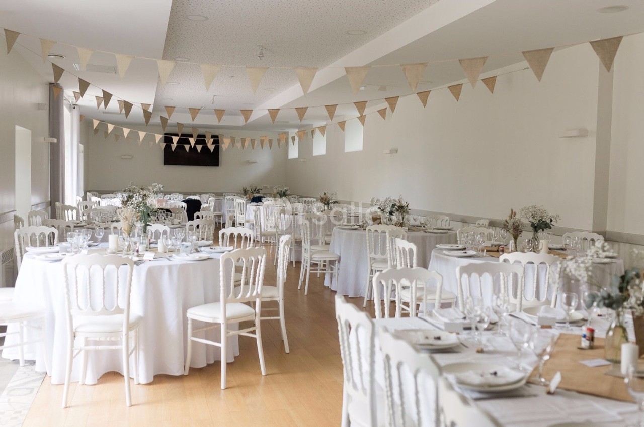Salle de réception décorée avec des tables rondes dressées, des chaises blanches et des guirlandes de fanions suspendues.