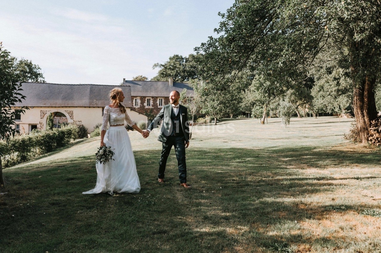Un couple en tenue de mariage marche main dans la main dans un jardin verdoyant près d'un bâtiment en pierre.