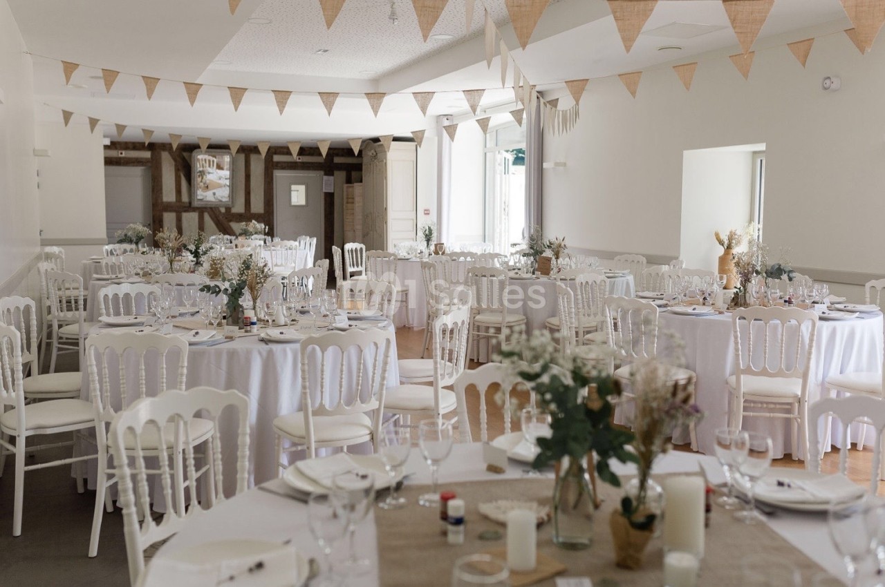 Salle de réception décorée pour un événement, avec des tables rondes dressées et des guirlandes en tissu suspendues.