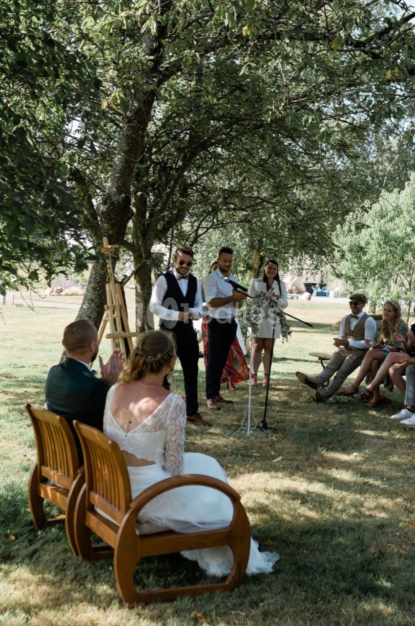 Un couple assis écoute un discours lors d'une cérémonie en plein air sous des arbres, entouré d'invités.