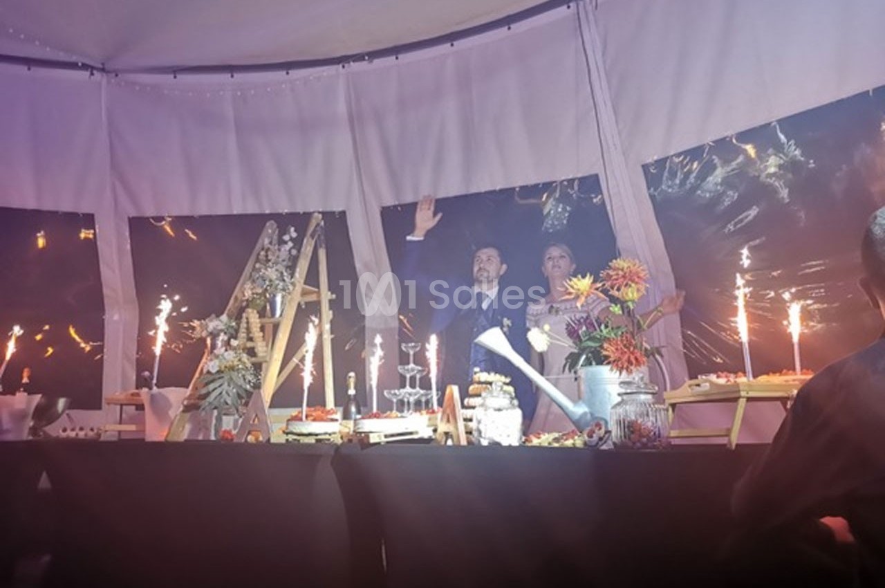 Un couple debout derrière une table décorée avec des desserts et des fleurs, dans une tente éclairée.