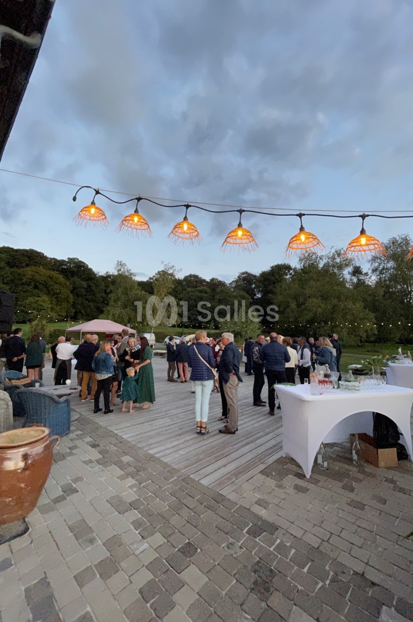Groupe de personnes réunies sur une terrasse extérieure en soirée, sous des lampes suspendues, avec un jardin en arrière…