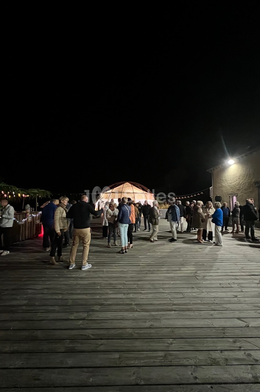 Groupe de personnes rassemblées sur une terrasse en bois la nuit, près d'une structure lumineuse et d'un bâtiment.