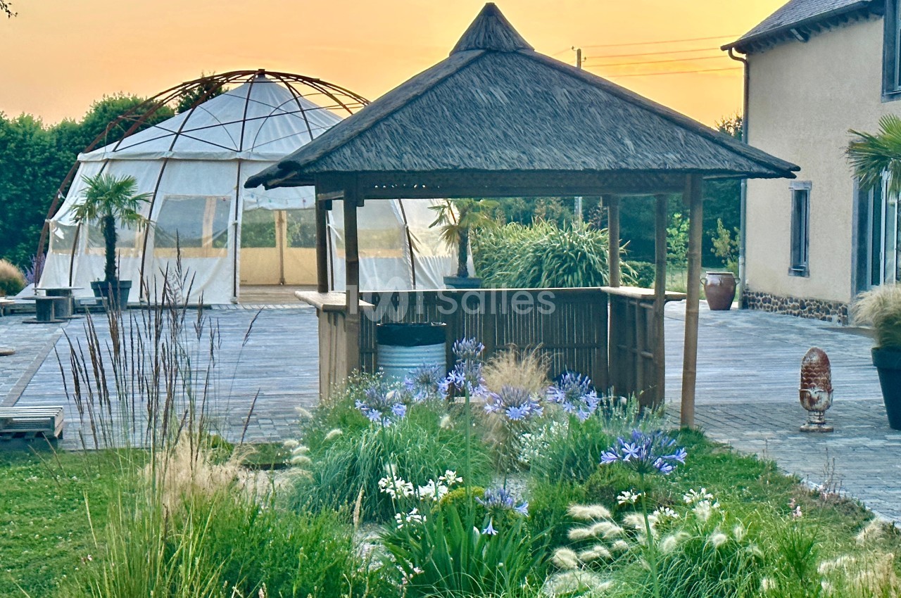 Kiosque en bois avec toit en chaume entouré de fleurs, près d'une terrasse et d'une structure en dôme, au coucher du soleil.