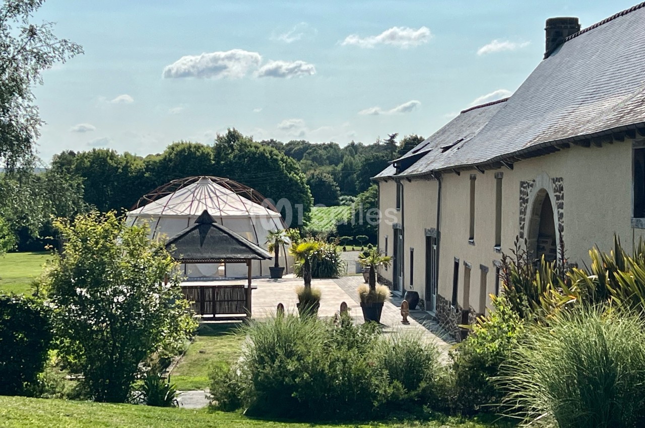 Bâtisse en pierre avec toit en ardoise, terrasse aménagée et structure géodésique dans un jardin verdoyant.
