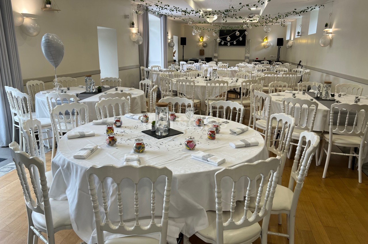 Salle de réception décorée avec des tables rondes dressées, nappes blanches, chaises blanches et guirlandes suspendues.