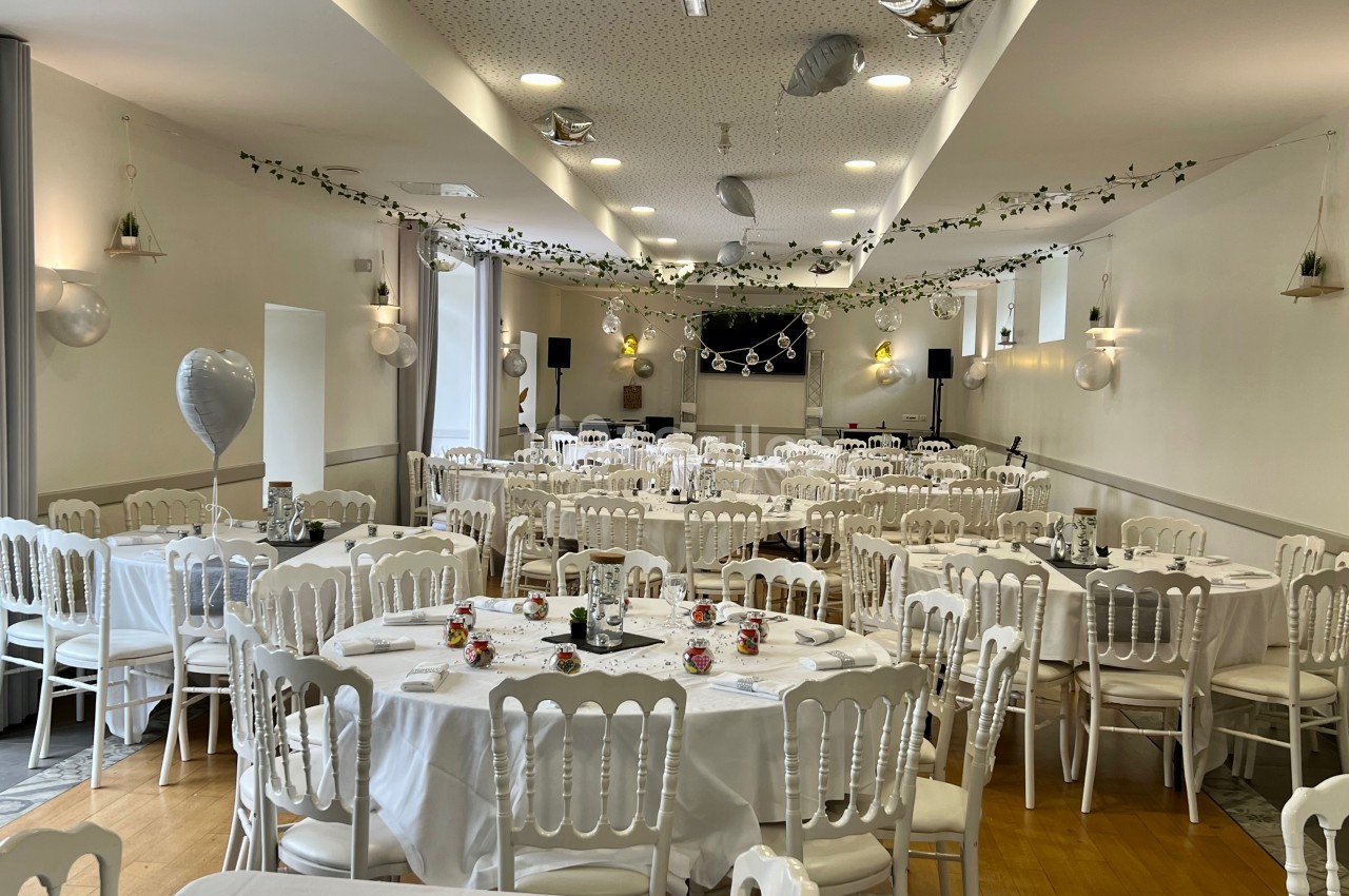 Salle de réception décorée avec des tables rondes, nappes blanches, chaises assorties et guirlandes suspendues.