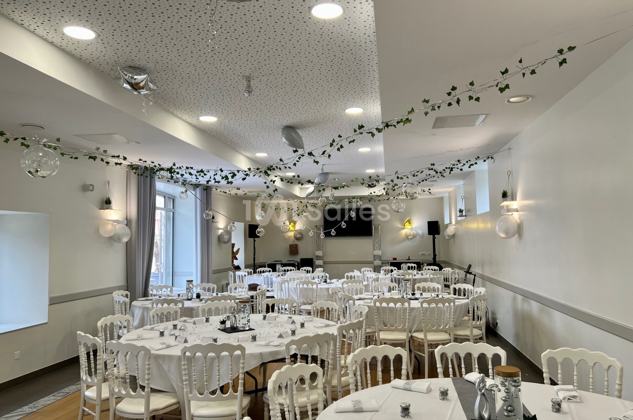Salle de réception décorée avec des guirlandes végétales, des tables rondes dressées et des chaises blanches.