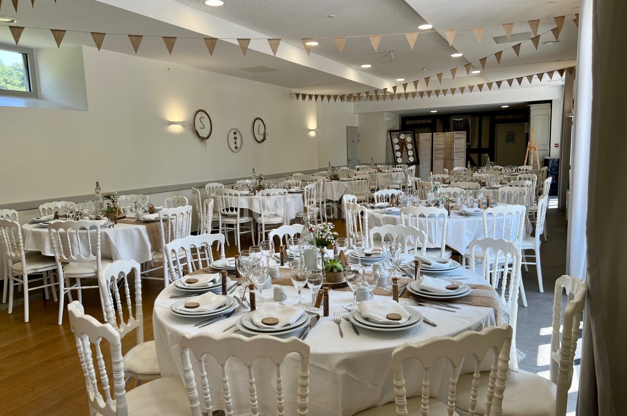Salle de réception décorée pour un événement, avec tables rondes dressées, chaises blanches et guirlandes triangulaires.