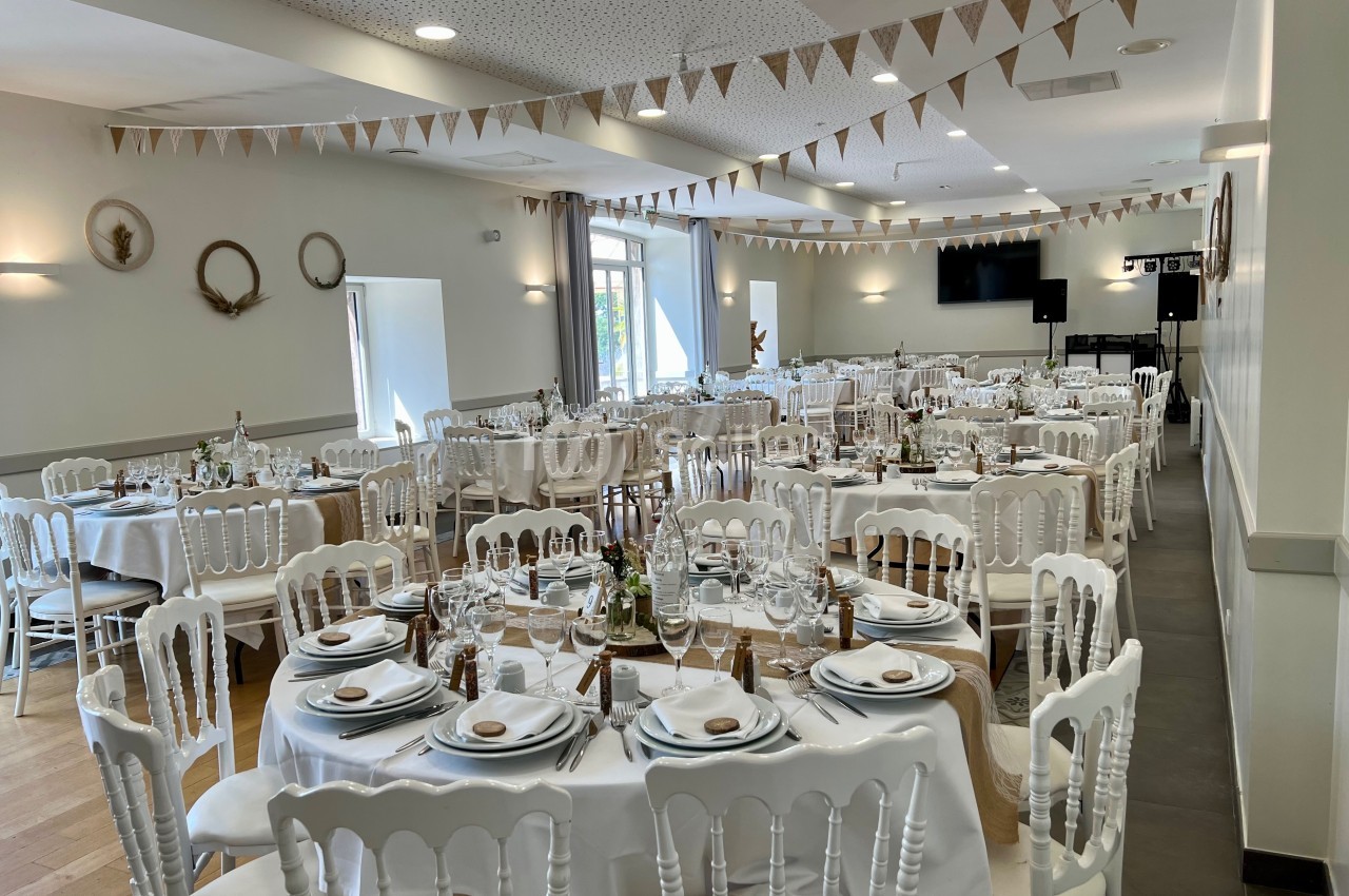Salle de réception décorée avec des tables rondes dressées, des chaises blanches et des guirlandes en tissu suspendues.