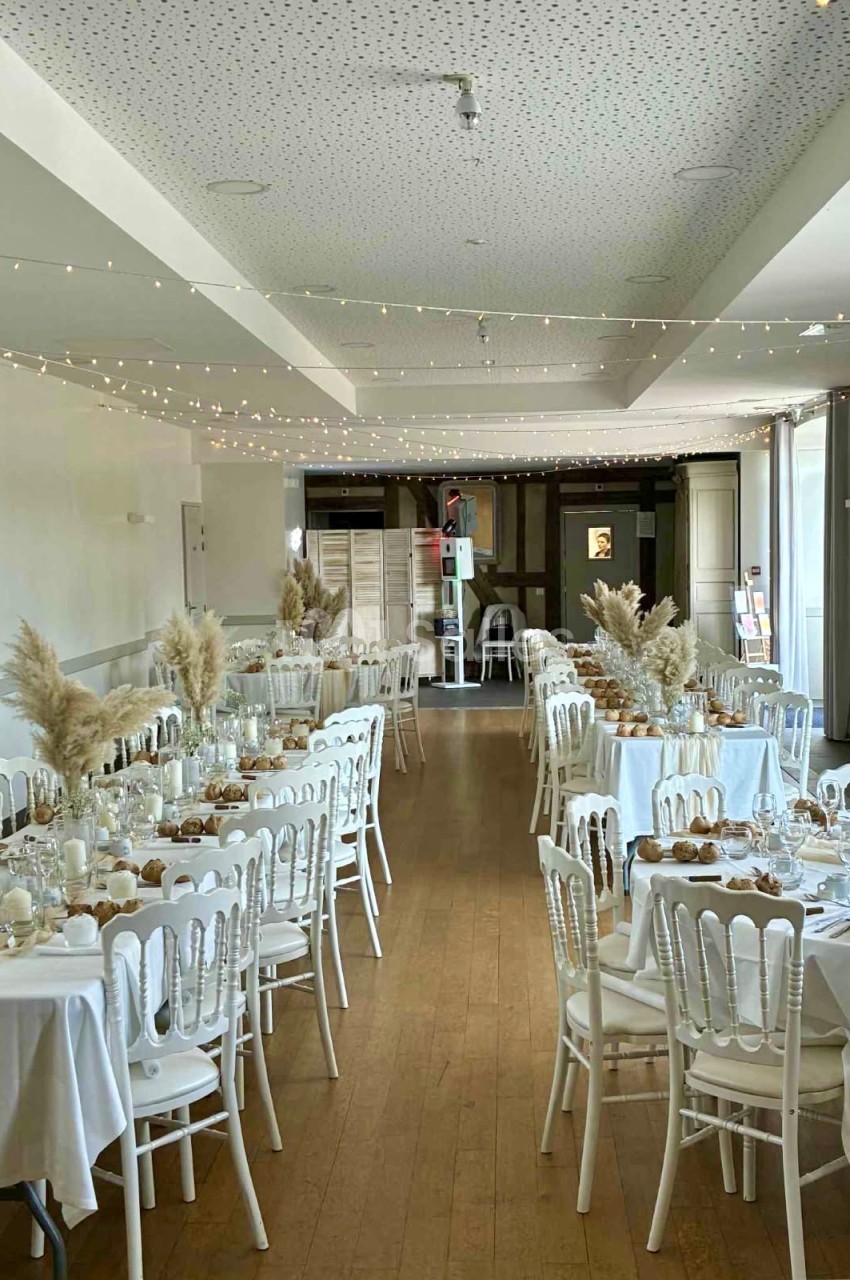 Salle de réception décorée avec des tables rondes, nappes blanches, chaises blanches et ornements de fleurs séchées.