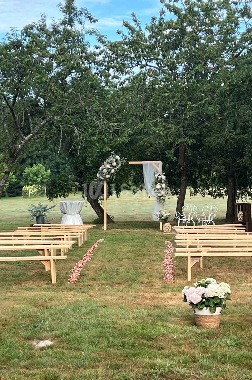 Cérémonie en plein air avec bancs en bois, arche fleurie et décor champêtre dans un jardin verdoyant.
