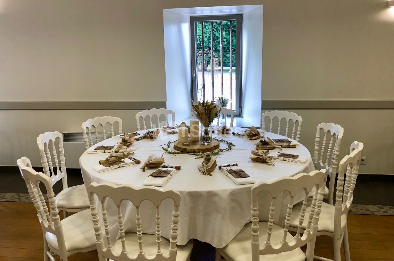 Table ronde décorée pour un événement, avec nappes blanches, chaises assorties et centre de table champêtre.