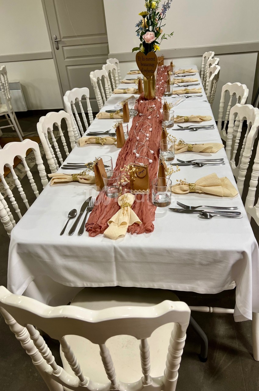 Table décorée pour un repas, avec nappe blanche, chemin de table rose, vaisselle, serviettes pliées et chaises blanches.