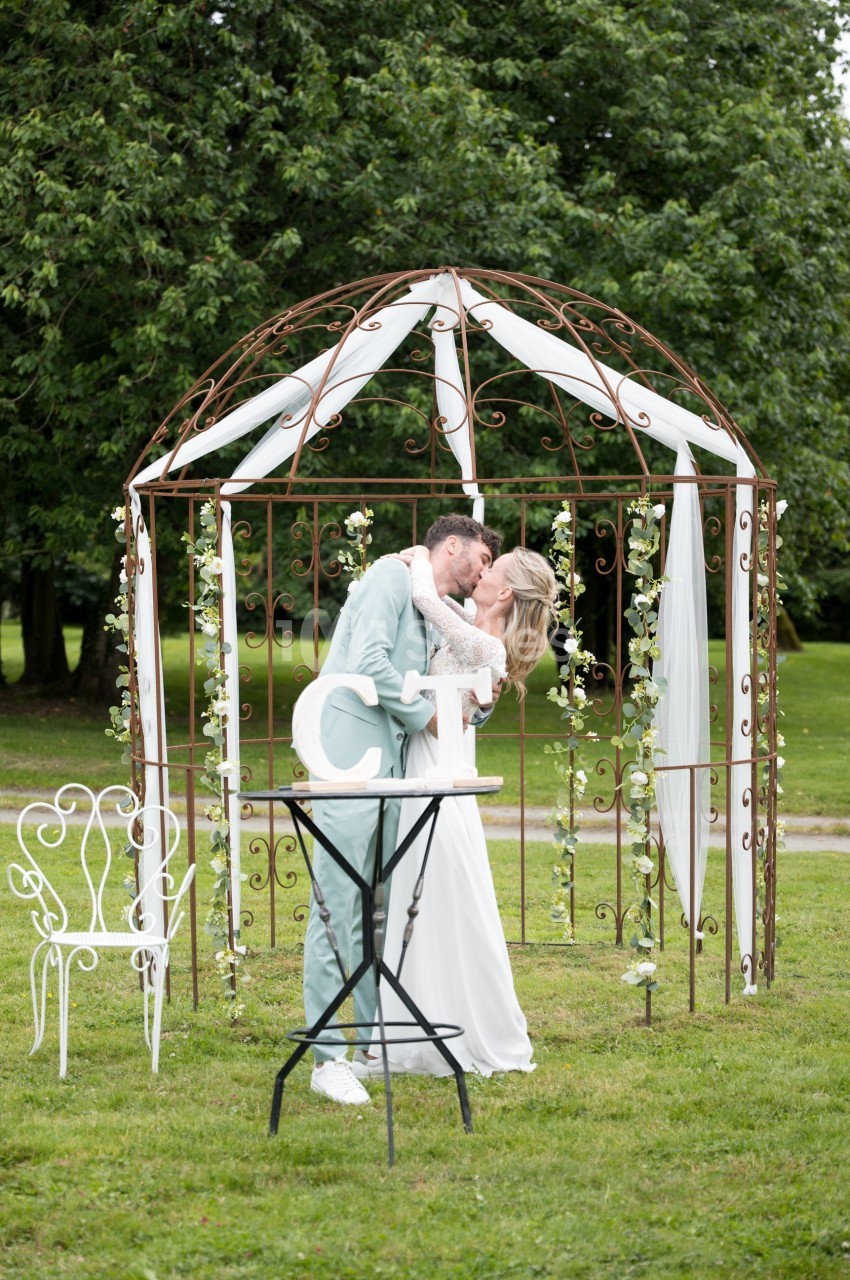 Un couple en tenue de mariage s'embrasse sous une pergola décorée de fleurs dans un jardin verdoyant.