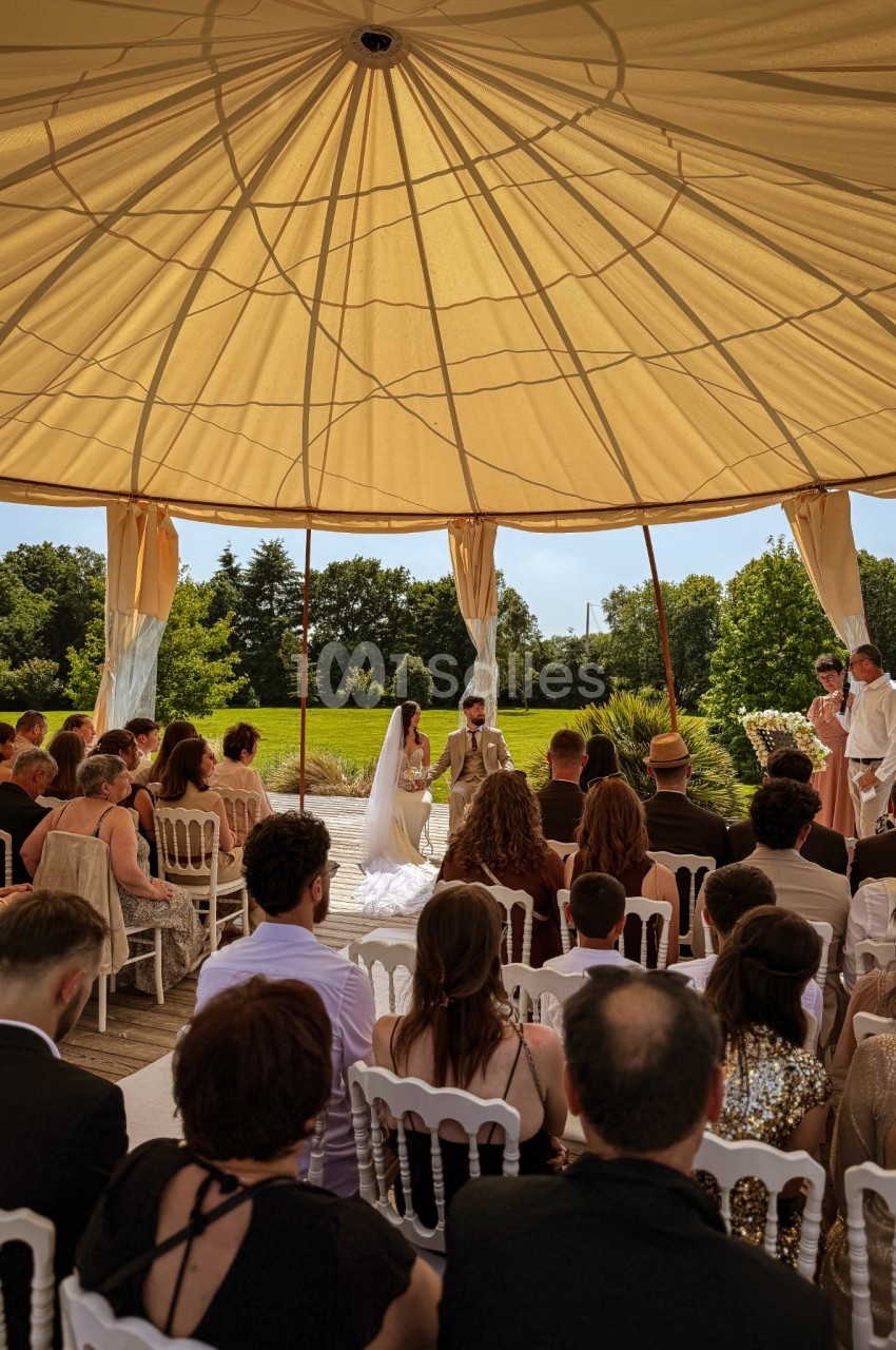 Cérémonie de mariage en extérieur sous une tente, avec les mariés face à l'officiant et des invités assis.
