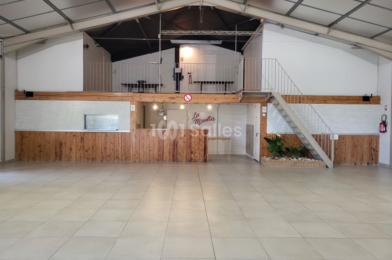 Salle spacieuse avec sol carrelé, mezzanine accessible par un escalier et mur en bois clair.