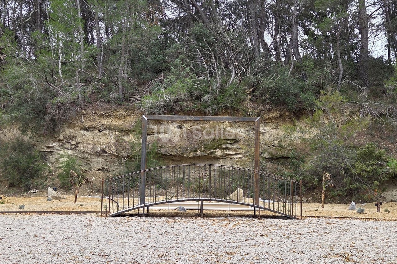 Petit pont métallique arqué traversant un espace gravillonné, avec une falaise et des arbres en arrière-plan.