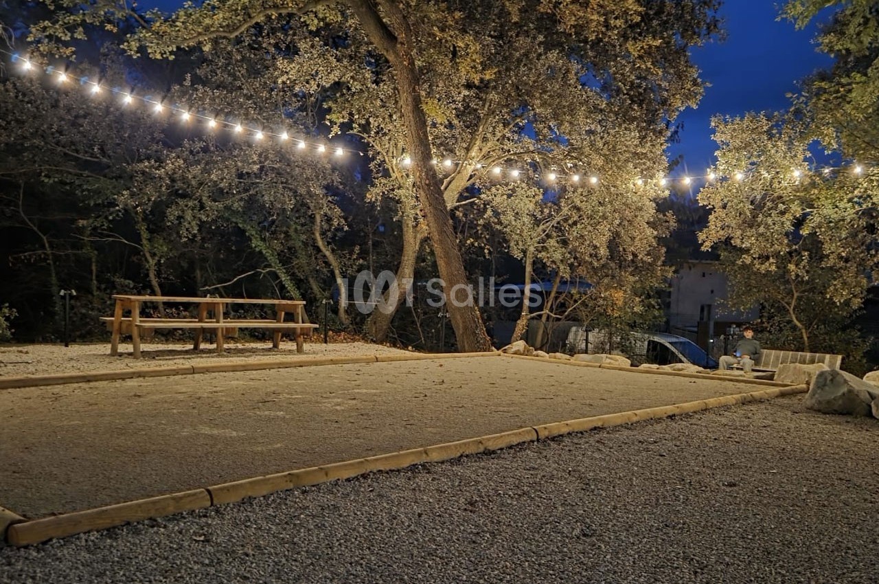 Terrain de pétanque en gravier éclairé par des guirlandes lumineuses, entouré d'arbres et de bancs en bois.