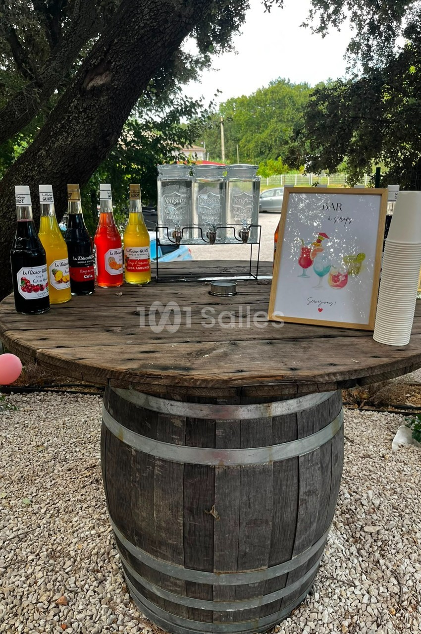 Table en tonneau avec sirops colorés, distributeurs d'eau, gobelets et affiche, installée en extérieur sous des arbres.