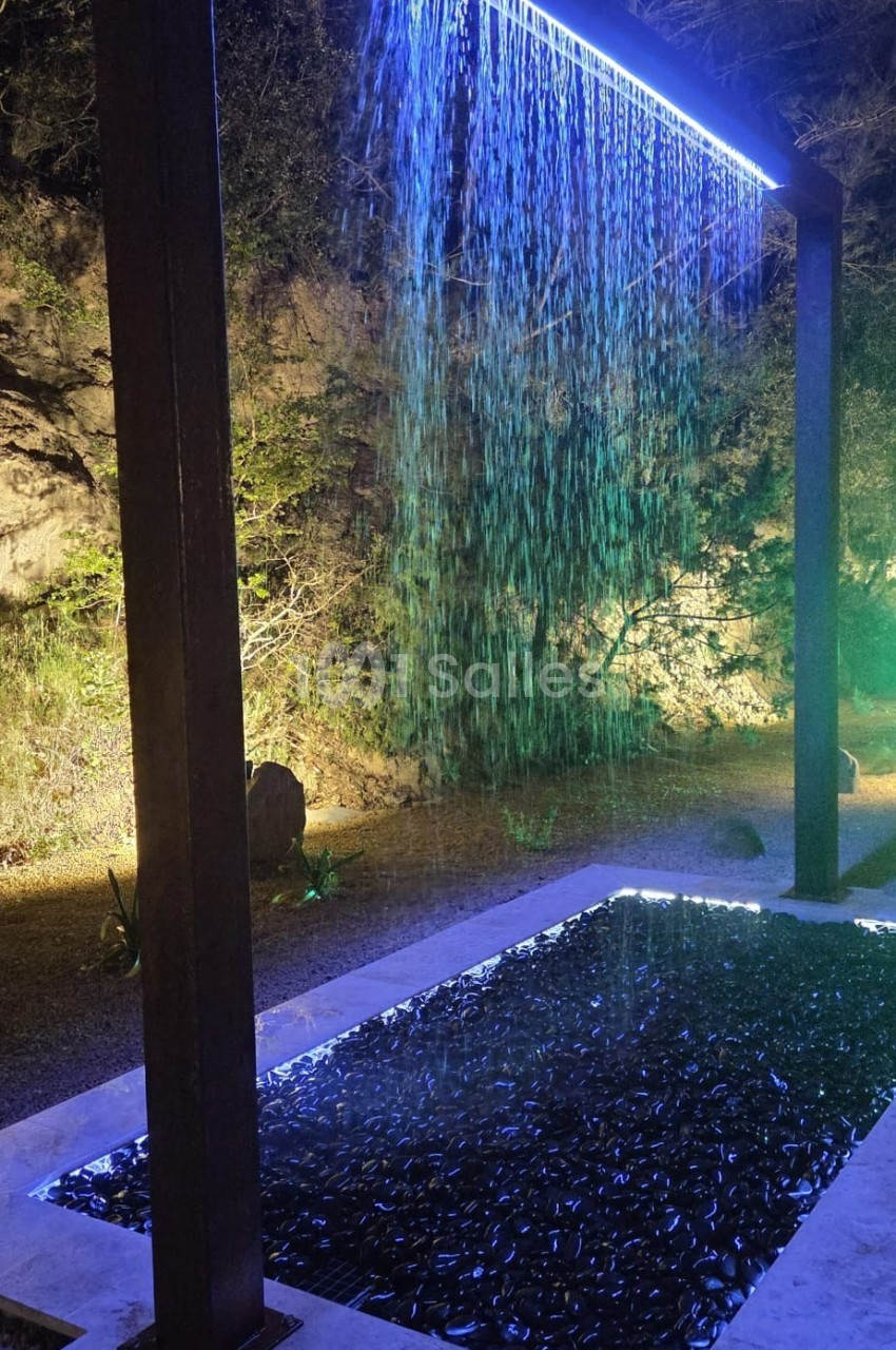 Cascade lumineuse artificielle avec éclairage bleu, tombant dans un bassin entouré de pierres, en extérieur nocturne.