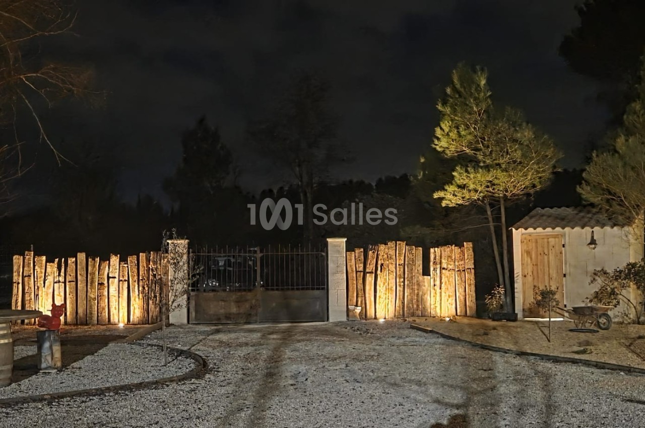 Entrée de propriété éclairée la nuit, avec portail en métal, clôture en bois et petite cabane sur la droite.