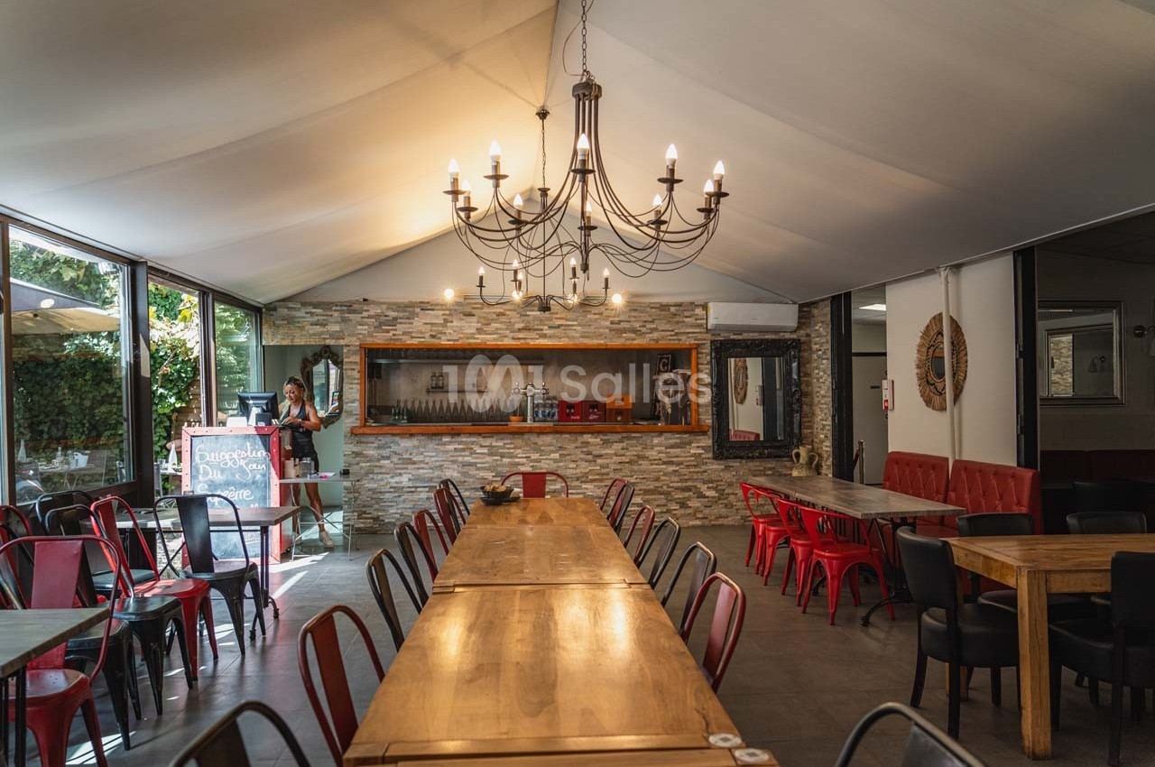 Salle de restaurant lumineuse avec de longues tables en bois, des chaises colorées et un lustre central.