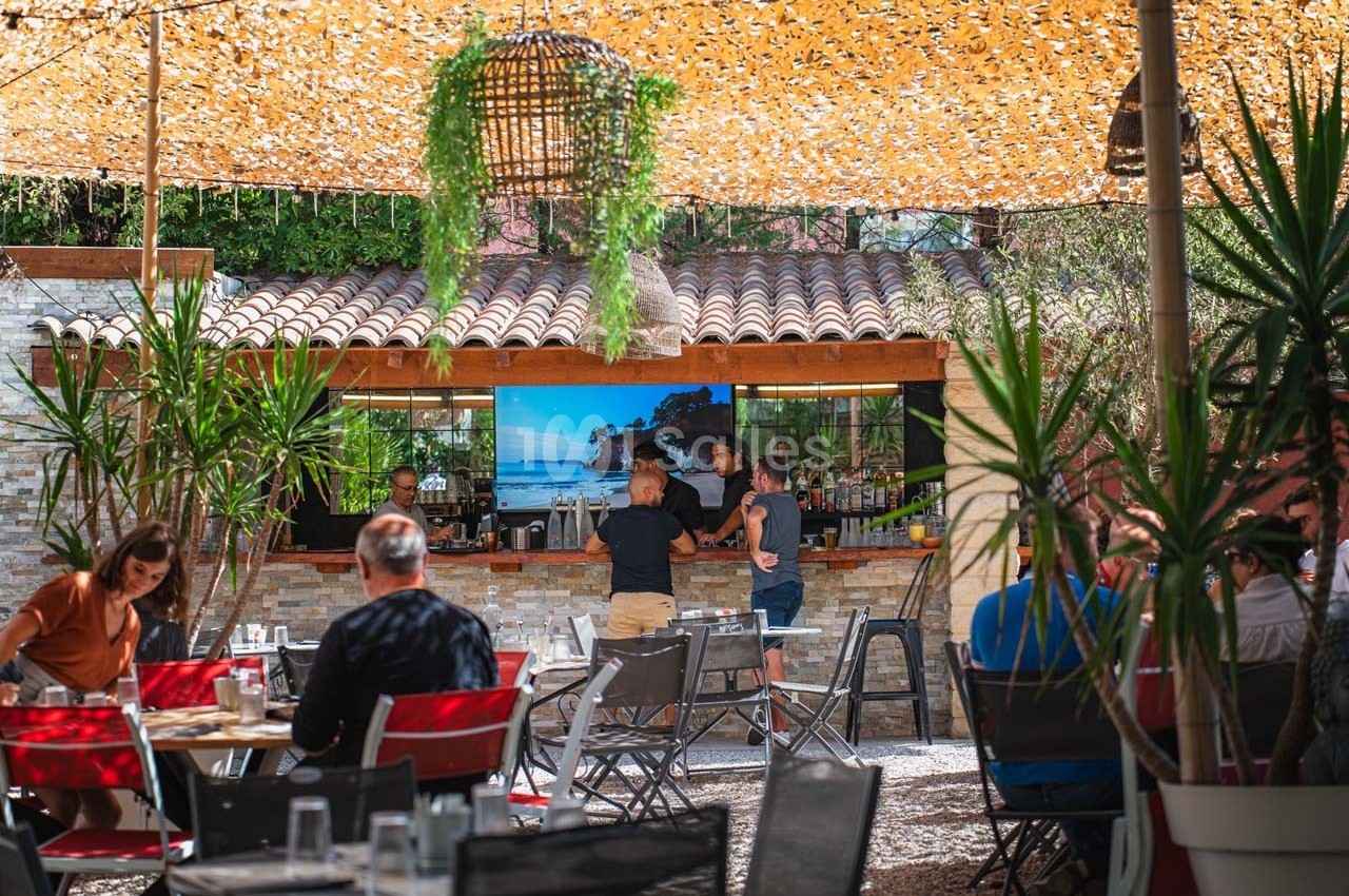 Terrasse ombragée avec des tables occupées, un bar en arrière-plan et des plantes décoratives autour.