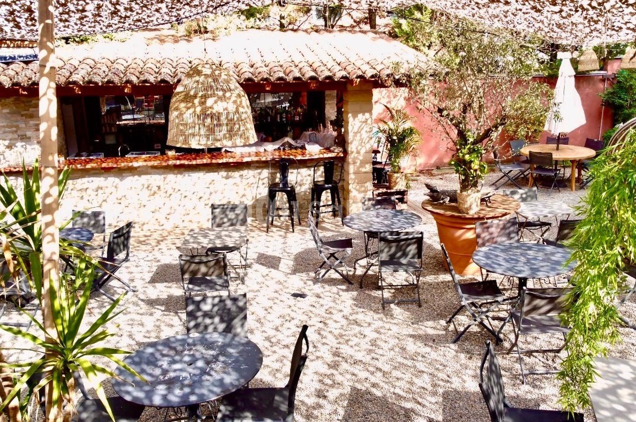 Terrasse extérieure ombragée avec des tables et chaises noires, entourée de végétation et d'un bar en arrière-plan.