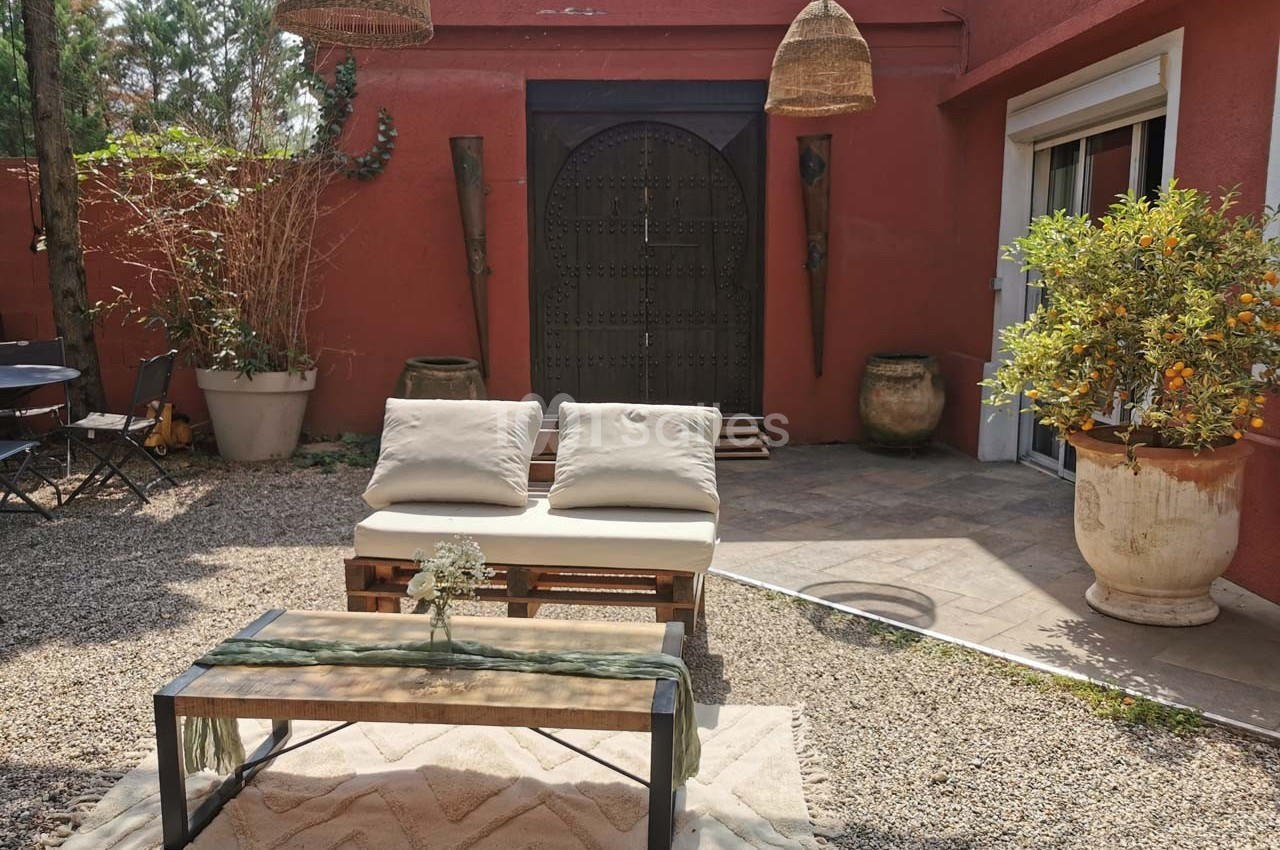 Salon de jardin extérieur avec canapé en palettes, table basse et plantes, devant une façade rouge avec porte en bois.